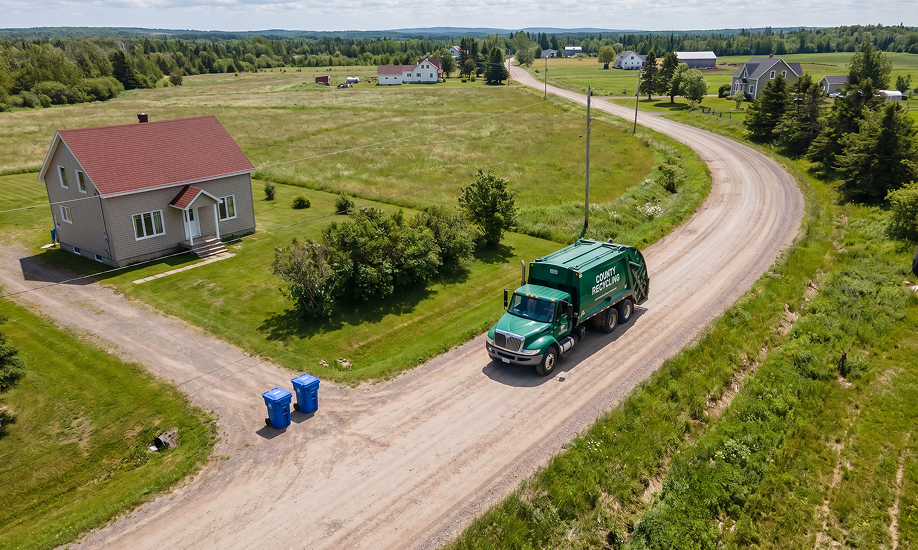Aerial view of a green recycling truck on a rural road near a house with blue bins.