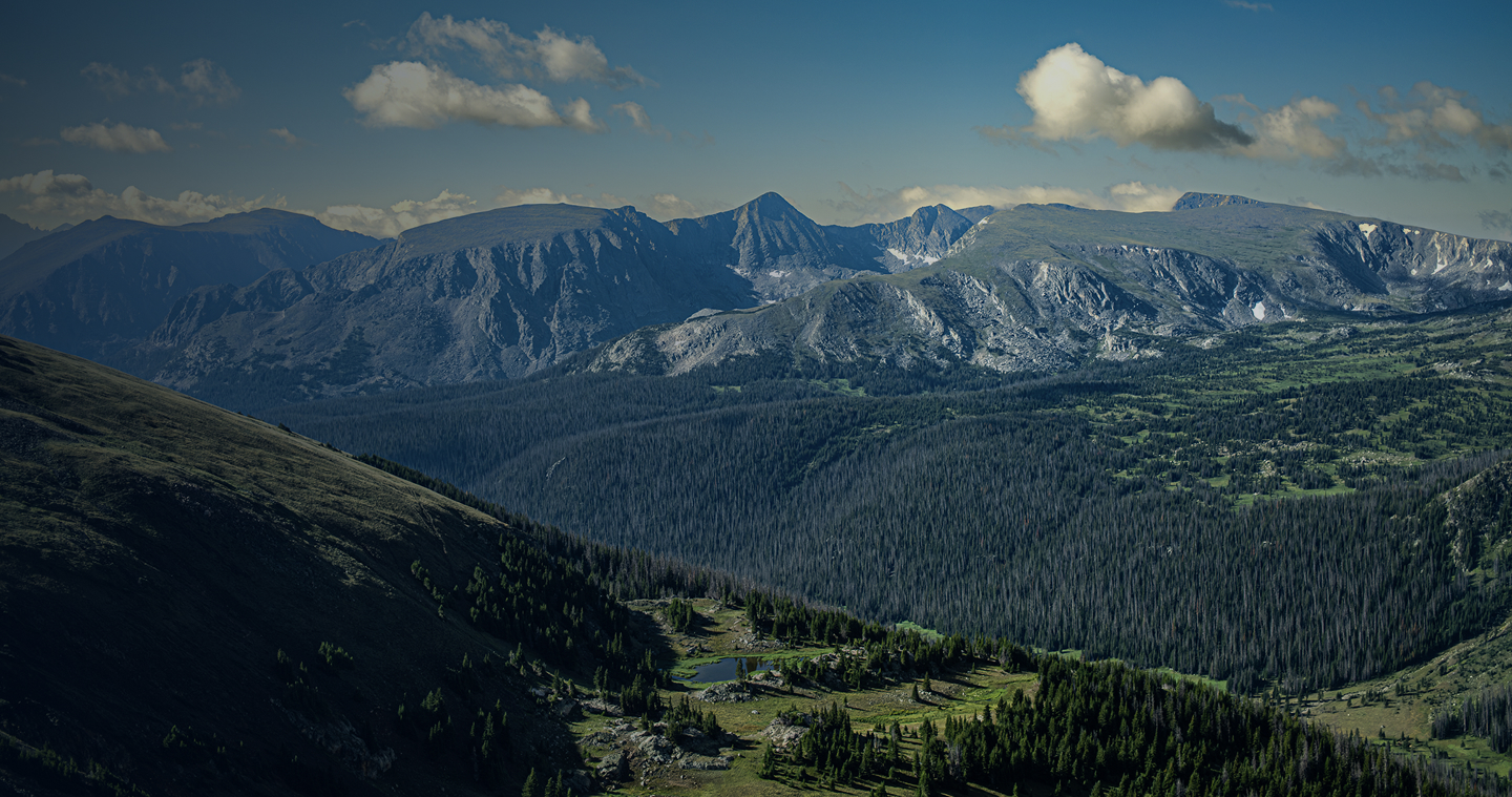 Panoramic view of a pine forest valley and alpine lake surrounded by rugged mountain peaks.