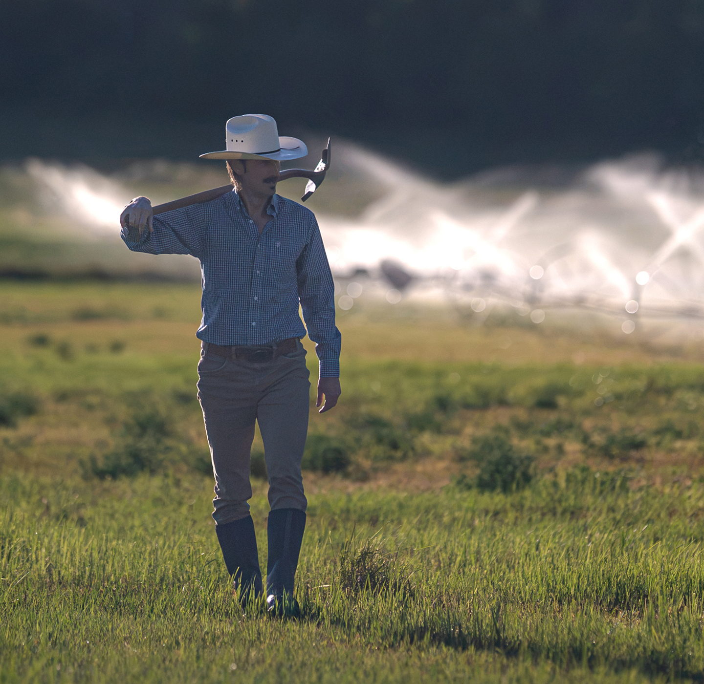 A farmer in a cowboy hat carrying a shovel across a green field with active irrigation sprinklers.