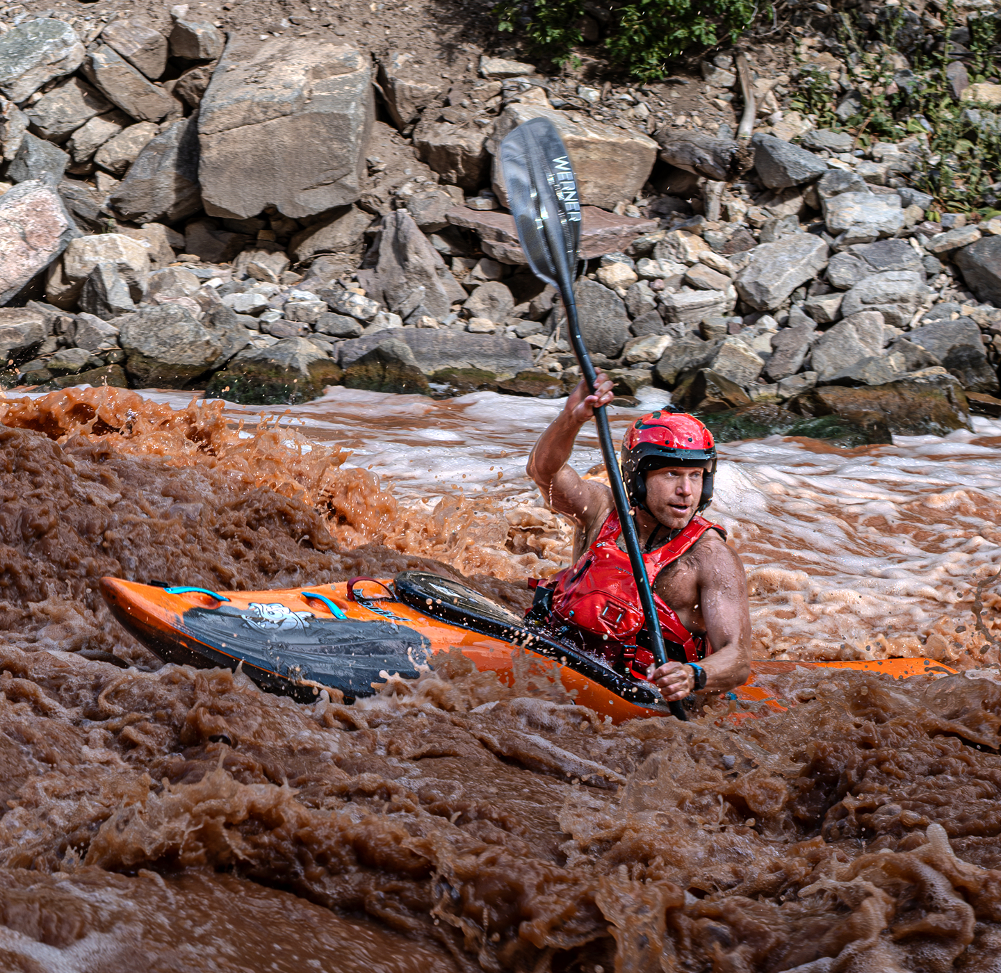 A kayaker in an orange kayak navigating muddy whitewater river rapids.