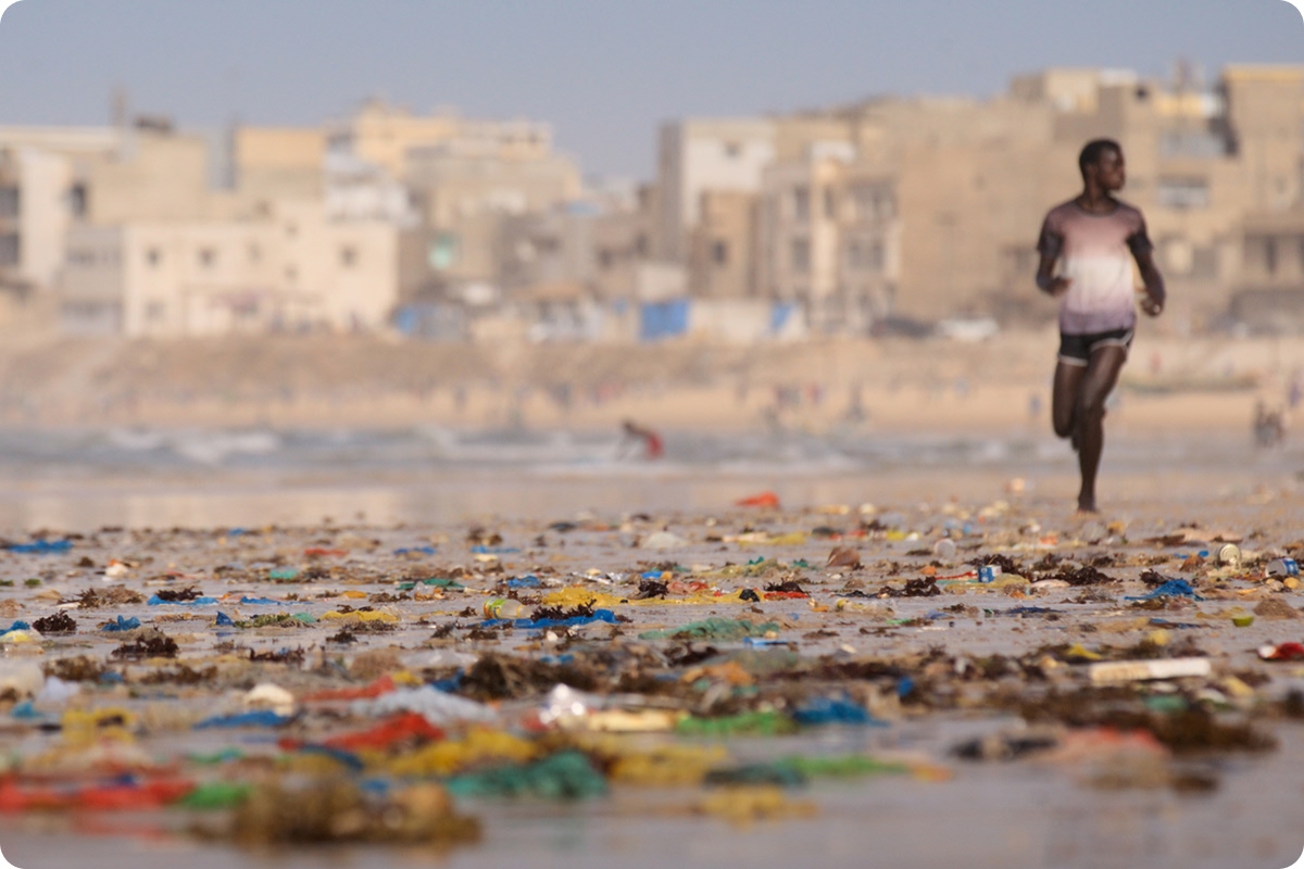 Man running on a beach littered with plastic waste in Senegal with buildings visible in the background.