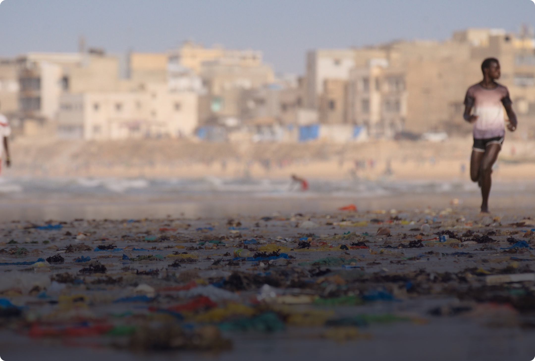 Man running on a beach littered with plastic waste in Senegal with buildings visible in the background.