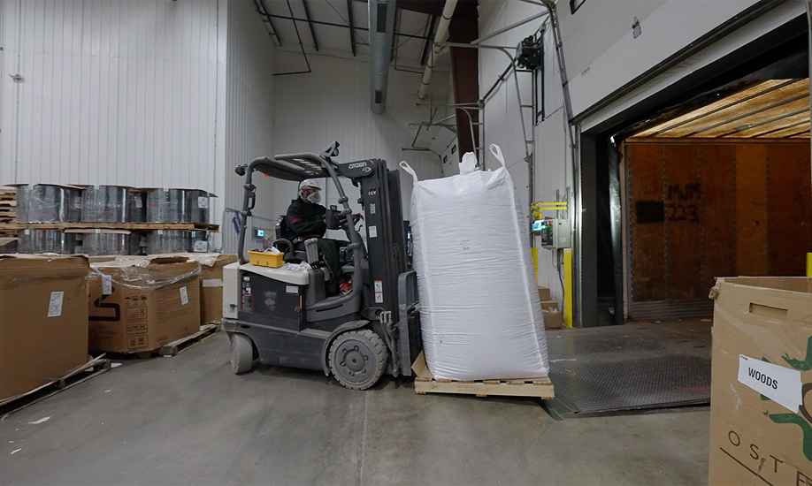 A warehouse worker wearing a hard hat and face mask operating a forklift to move a large white bulk bag on a wooden pallet near an open loading dock door.