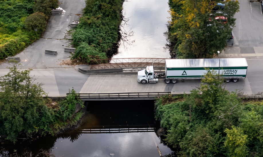 An aerial view of a white semi-truck pulling a green and white trailer with the text URBAN IMPACT as it drives across a concrete bridge over a tree-lined river.