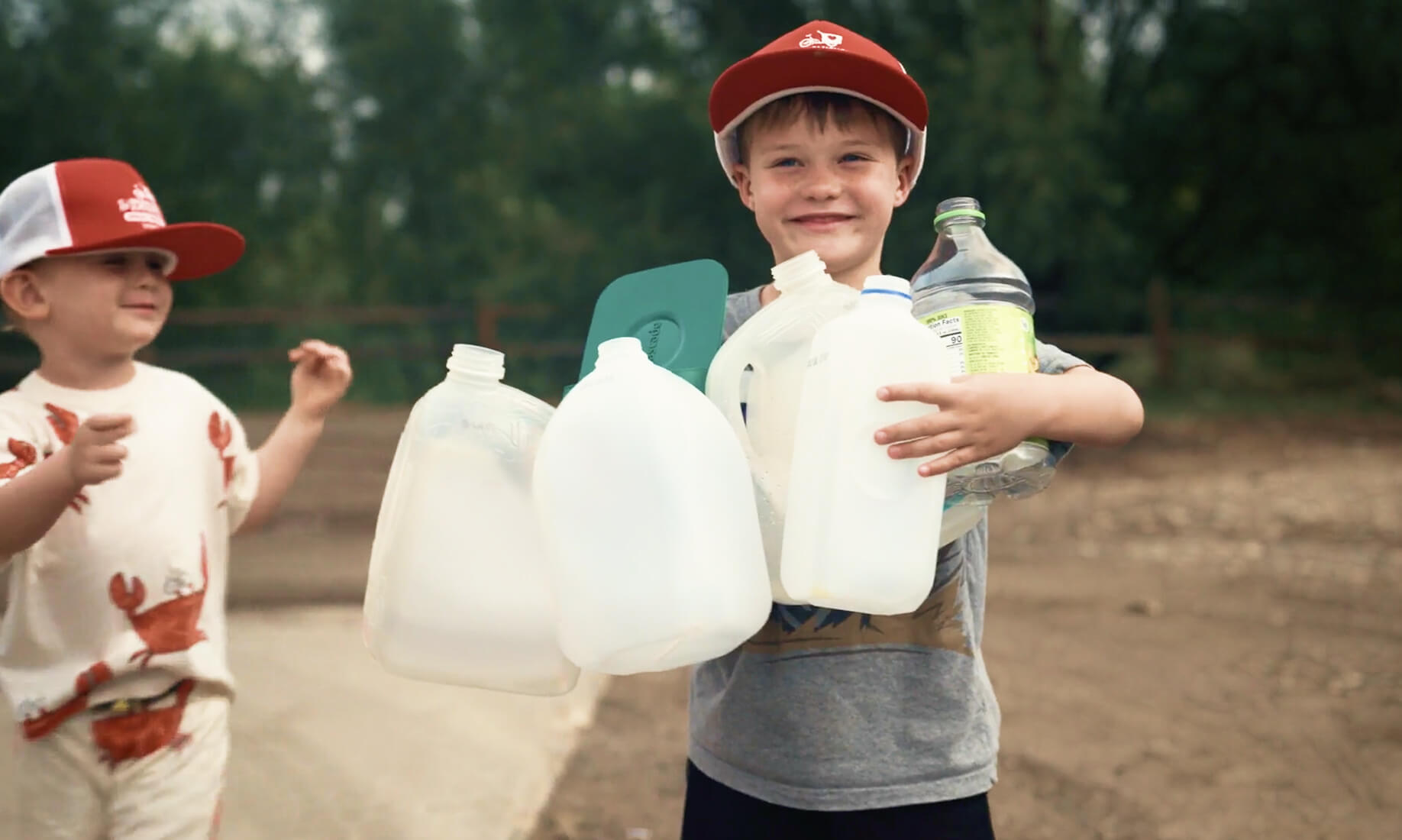 A smiling young boy wearing a red baseball cap holding a large bundle of empty plastic milk jugs and bottles outdoors, with a younger child in a matching cap standing beside him.